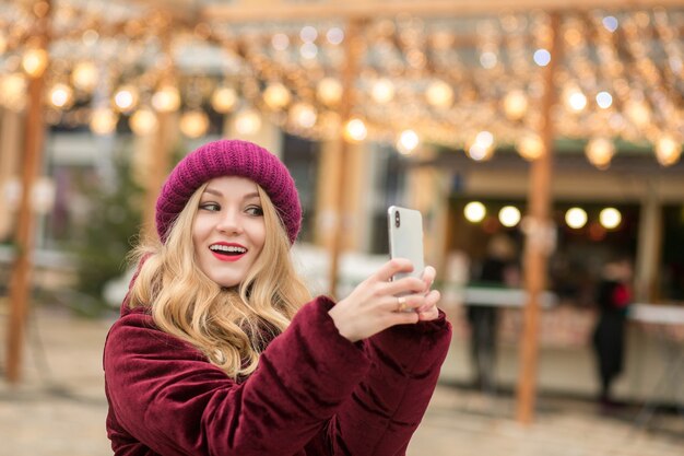 Linda mulher loira vestida com roupas quentes fazendo selfie no fundo de uma guirlanda em Kiev