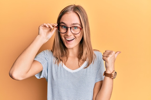 Foto linda mulher loira vestindo roupas casuais e segurando os óculos apontando o polegar para o lado sorrindo feliz com a boca aberta