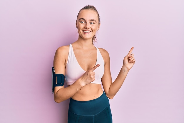 Linda mulher loira vestindo roupas esportivas e faixa de braço sorrindo e olhando para a câmera apontando com as duas mãos e dedos para o lado