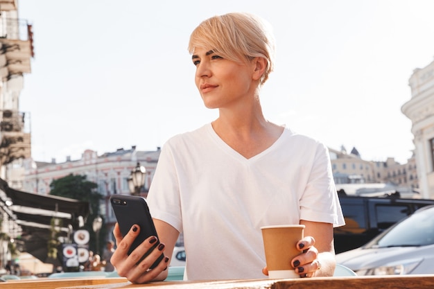 linda mulher loira vestindo uma camiseta branca usando um telefone celular, enquanto está sentada em um café ou restaurante no verão e bebendo café em um copo de papel
