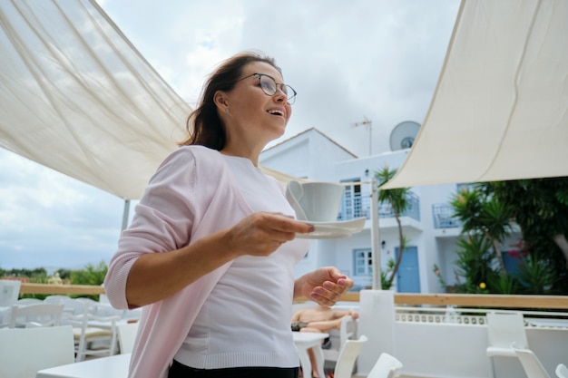 Linda mulher madura, aproveitando as férias no hotel resort, no restaurante, vista da paisagem montanhosa. Mulher andando com uma xícara de café. Lazer, verão, fim de semana, turismo, viagens pessoas de meia-idade