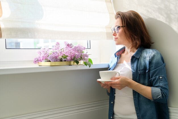 Linda mulher madura em casa com flores, sorrindo feminino em pé perto da janela com uma xícara de chá