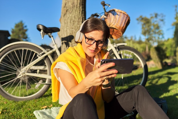 Linda mulher madura em fones de ouvido com smartphone falando online, comunicação por vídeo. Feliz mulher de meia idade fazendo videochamada relaxando sentada no parque, fundo de dia ensolarado de bicicleta