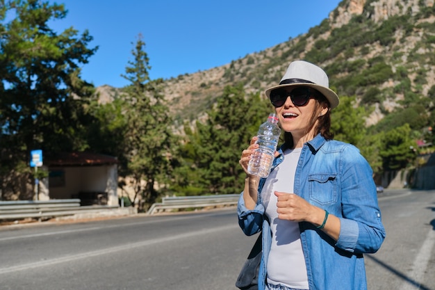 Linda mulher madura em pé na estrada da montanha bebendo água da garrafa em um dia quente de sol. Agindo estilo de vida saudável e alimentação saudável