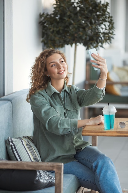 Linda mulher milenar com cabelo encaracolado sentado no café da rua e fazendo selfie no telefone