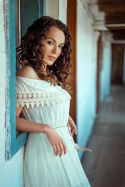 Linda mulher morena cacheada caucasiana usando um vestido branco e encostada na janela