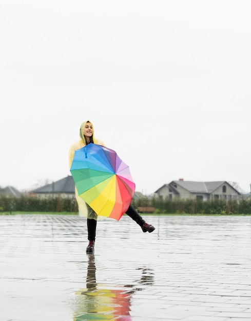 Linda mulher morena com capa de chuva amarela segurando guarda-chuva arco-íris na chuva