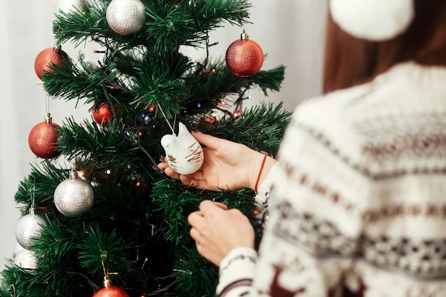 Linda mulher morena com suéter de inverno de rena e lindo chapéu de Papai Noel vermelho sorrindo e decorando a árvore de Natal verde com enfeites vermelhos e prateados e conceito de cartão de saudação de luzes