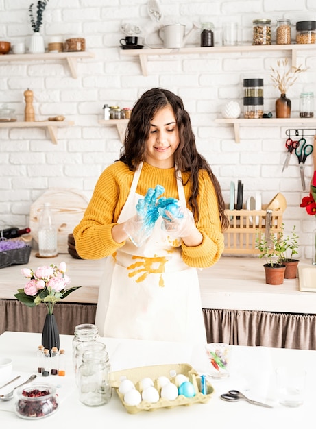 Linda mulher morena com um suéter amarelo e um avental cinza para colorir ovos de Páscoa de azul na cozinha