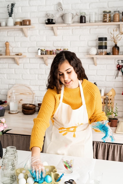 Linda mulher morena com um suéter amarelo e um avental cinza para colorir ovos de Páscoa de azul na cozinha