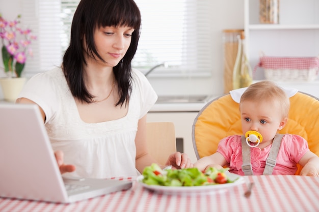 Linda mulher morena comendo uma salada ao lado de seu bebê enquanto relaxa com seu laptop