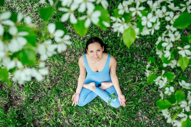 Linda mulher morena em um terno azul desportivo num contexto de árvores floridas de primavera, sentado na grama verde e fazendo relaxamento de ioga, o conceito de meditação.