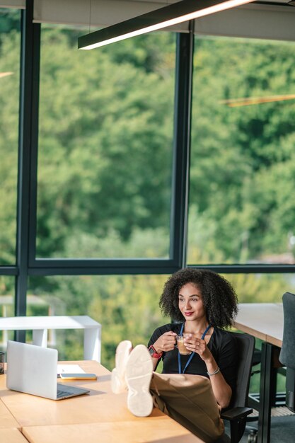 Linda mulher morena parecendo satisfeita enquanto está sentada no escritório e tomando café