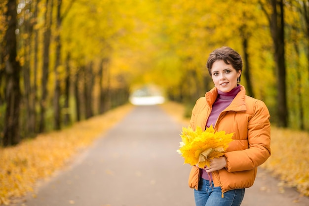 Linda mulher morena posando em uma jaqueta amarela em um parque de outono com um buquê de folhas de outono