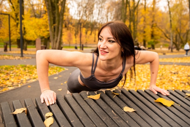 Linda mulher morena se aquecendo e fazendo algumas flexões no parque outono. Ela faz flexões no banco com folhas amarelas. Ela está sorrindo.