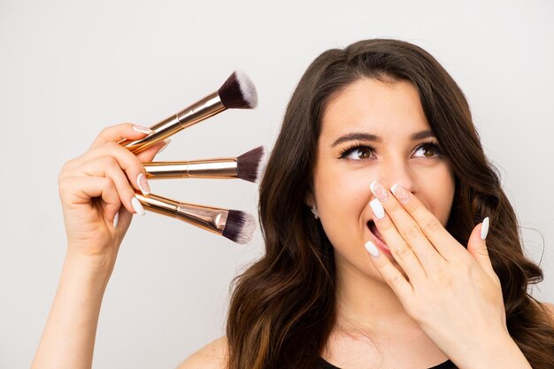 Linda mulher morena segurando pincéis de maquiagem cobrindo a boca com a mão