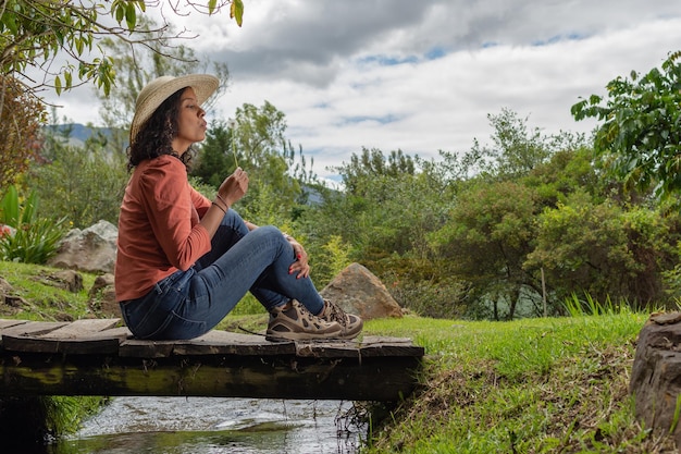 Linda mulher morena usando chapéu e roupas casuais, sentado em uma velha ponte de madeira sobre um pequeno riacho enquanto sopra algumas sementes de dente-de-leão