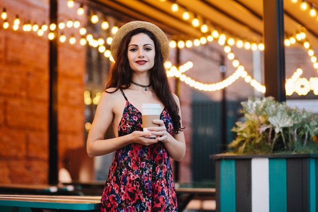 Linda mulher morena usando chapéu e vestido em pé na cafeteria segurando o copo de papel com café ou chá Jovem empresária jantando no café descansando depois do trabalho duro no escritório Conceito de beleza