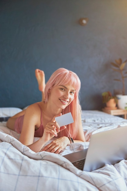 Linda mulher mostra um cartão de crédito em branco perto de um laptop moderno em uma cama grande