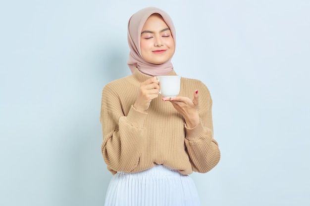 Linda mulher muçulmana asiática de suéter marrom segurando caneca e bebendo chá isolado sobre fundo branco Conceito de estilo de vida religioso de pessoas