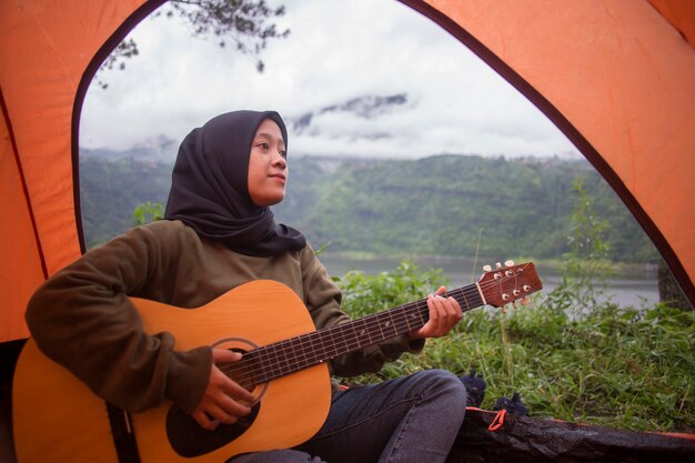 Linda mulher muçulmana asiática tocando guitarra sentada perto do lago na montanha com vista para o rio com mochila, barraca ao pôr do sol