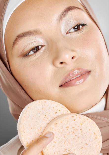 Linda mulher muçulmana segurando e usando esponjas de maquiagem no rosto contra um fundo cinza do estúdio Jovem mulher vestindo um hijab e fazendo sua rotina de cuidados com a pele cuidando da pele saudável