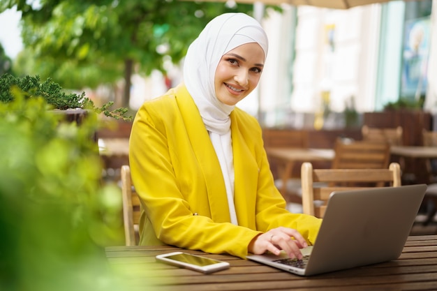 Linda mulher muçulmana sorridente com laptop no café