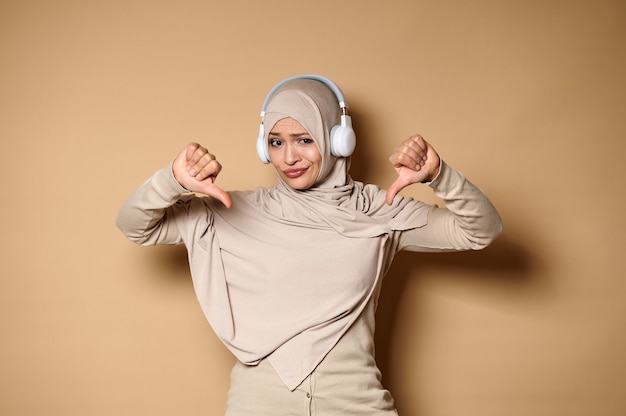 Linda mulher muçulmana usando hijab e fones de ouvido mostrando os polegares para baixo para a câmera, em pé contra bege.