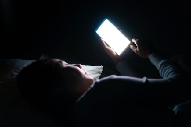 Linda mulher na cama usando smartphone à noite, no quarto escuro. Conceito de dependência de telefone celular