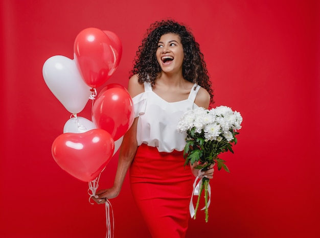 Linda mulher negra com balões em forma de coração e buquê de flores brancas, isoladas no vermelho