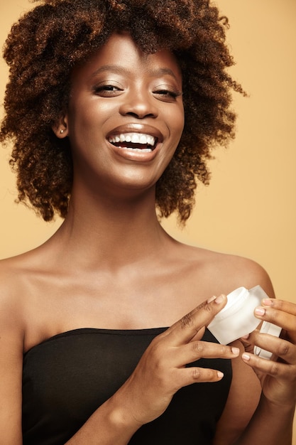 Foto linda mulher negra com pele limpa saudável e cabelos cacheados em estilo afro segurando pote de creme corporal