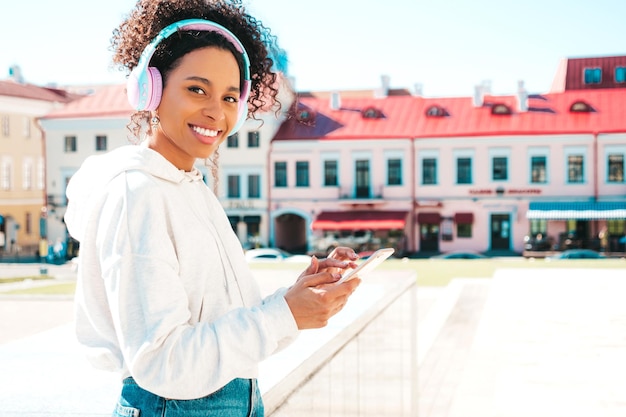 Linda mulher negra com penteado de cachos afro Modelo sorridente com capuz branco Mulher despreocupada sexy curtindo ouvir música em fones de ouvido sem fio Posando no fundo da rua ao pôr do sol Segura o telefone