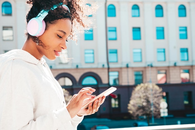 Linda mulher negra com penteado de cachos afro Modelo sorridente com capuz branco Mulher despreocupada sexy curtindo ouvir música em fones de ouvido sem fio Posando no fundo da rua ao pôr do sol Segura o telefone