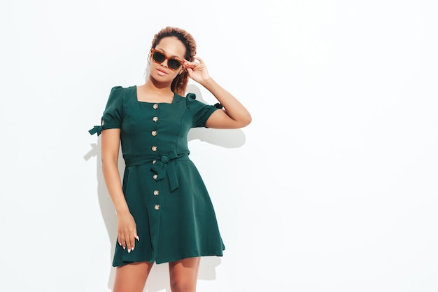 Linda mulher negra com penteado de cachos afro Modelo sorridente vestido com vestido branco de verão verde Mulher despreocupada sexy posando perto da parede no estúdio Bronzeado e alegre isolado em óculos de sol