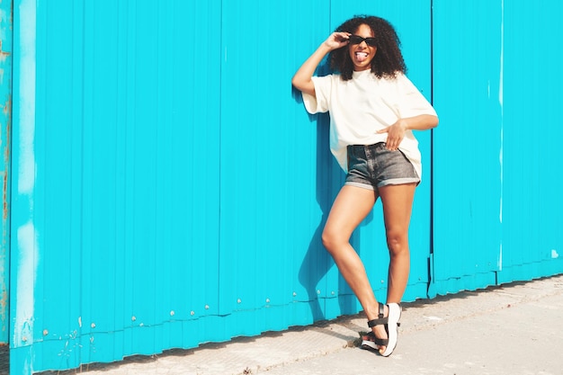 Foto linda mulher negra com penteado de cachos afromodelo de hipster sorridente em camiseta branca mulher despreocupada sexy posando na rua perto da parede azul em óculos de sol alegre e feliz mostra a língua