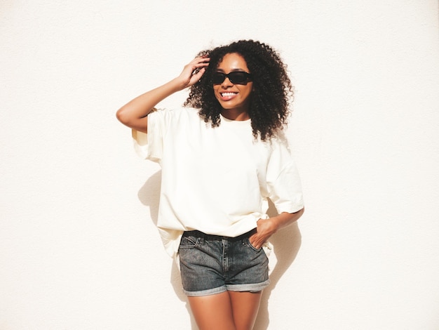 Foto linda mulher negra com penteado de cachos afromodelo de hipster sorridente em camiseta branca mulher despreocupada sexy posando na rua perto de parede branca em óculos de sol alegre e feliz