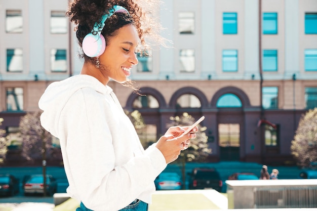 Linda mulher negra com penteado de cachos afroModelo sorridente com capuz amareloFêmea despreocupada sexy gostando de ouvir música em fones de ouvido sem fioPosando no fundo da rua ao pôr do sol Segura o telefone