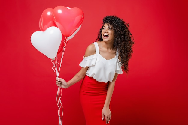 Linda mulher negra elegante segurando balões de hélio com sorriso feliz isolado no vermelho