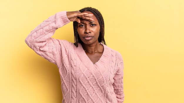 Linda mulher negra negra parecendo perplexa e atônita, com a mão na testa olhando para longe, observando ou procurando