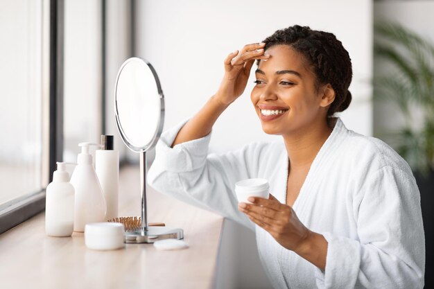 Linda mulher negra sentada perto do espelho e aplicando creme hidratante no rosto