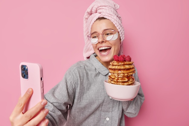 Linda mulher positiva ri e posa para selfie usa smartphone mostra delicioso café da manhã preparado panquecas apetitosas com framboesas vestidas com roupa de dormir passa por procedimentos de beleza em casa