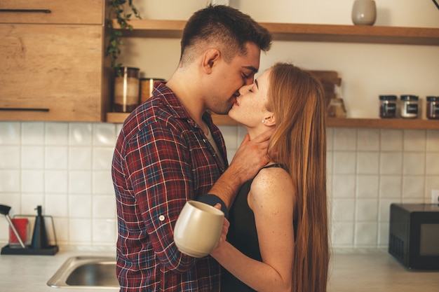 Linda mulher ruiva com sardas beijando o amante enquanto bebe uma xícara de chá na cozinha