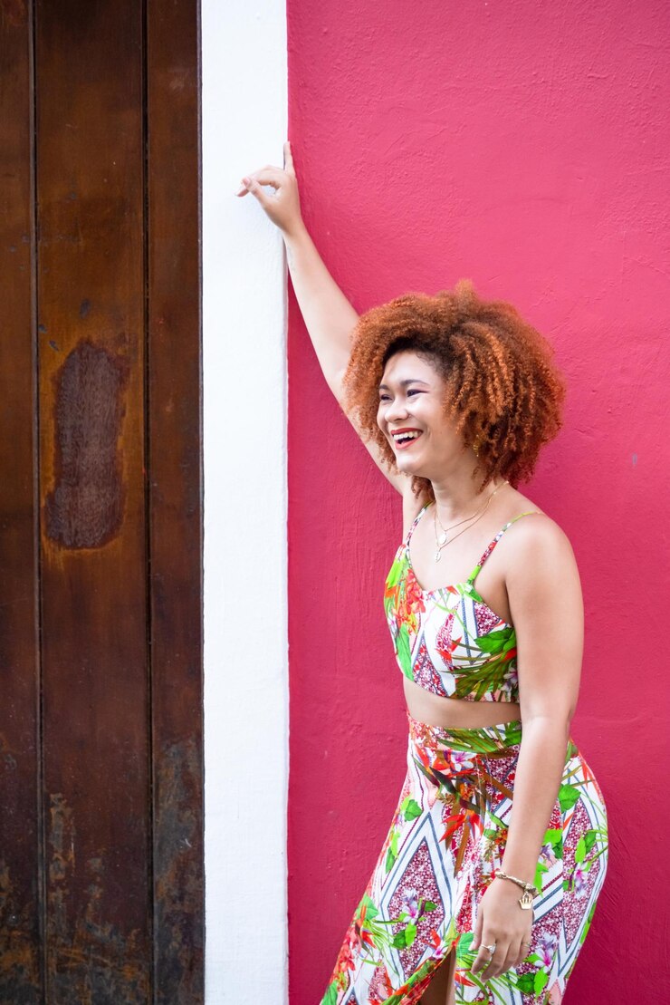 Foto linda mulher ruiva com vestido longo e colorido em pé contra uma parede vermelha pessoa feliz viajando