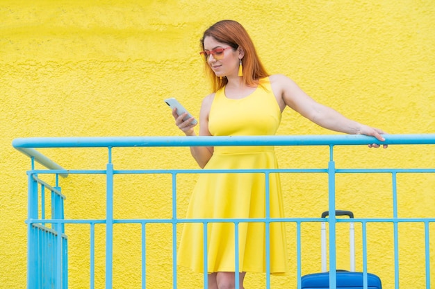 Linda mulher ruiva fica com uma mala azul e usa um smartphone em um fundo amarelo