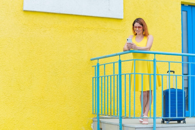 Linda mulher ruiva fica com uma mala azul e usa um smartphone em um fundo amarelo