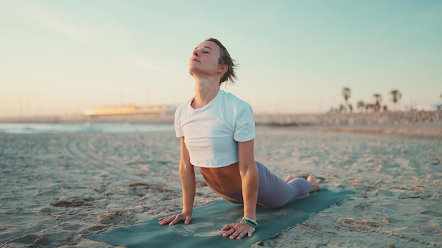 Linda mulher se exercitando no tapete de ioga ao ar livre Garota fitness em roupas esportivas praticando ioga na praia