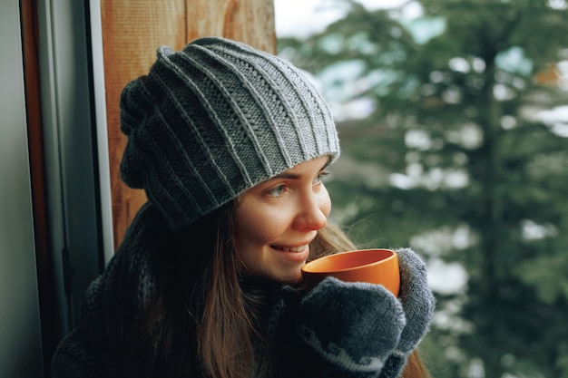 Linda mulher segurando e bebendo uma xícara de café ou chocolate em luvas sentado em casa perto da janela. fundo de árvore de neve de inverno turva.