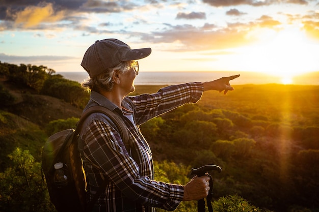 Linda mulher sênior de chapéu e roupas casuais caminhadas na natureza desfrutando de liberdade e beleza Fêmea madura feliz apontando um dedo para o raio de sol amarelo sobre a água sorrindo Horizonte sobre o mar