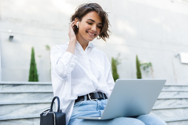 Linda mulher sentada ao ar livre usando um laptop ouvindo música com fones de ouvido