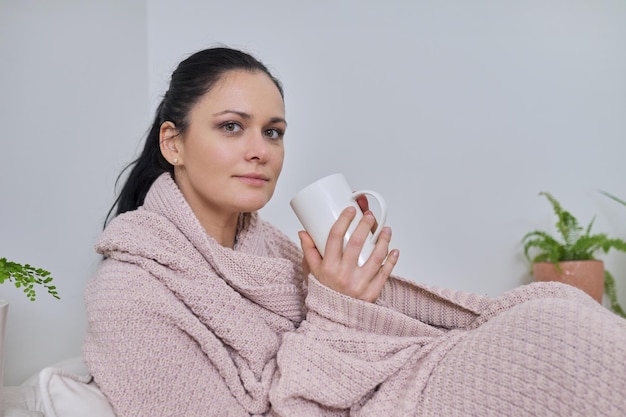 Linda mulher sentada em casa sob o cobertor quente de malha com uma xícara de chá